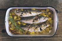 Roast mackerel with potatoes in a roasting dish over wooden surface — Stock Photo
