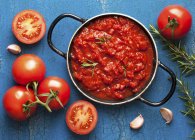 Tomatensauce mit Rosmarin im Topf, umgeben von frischen Zutaten über blauer Oberfläche — Stockfoto
