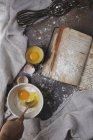 Top view of a baking scene with eggs, flour, a recipe book and a whisk — Stock Photo