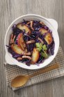 Red cabbage with apple and bacon in white dish over towel on wooden surface with spoon — Stock Photo