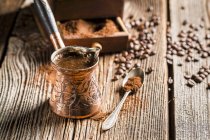 Крупный план турецкого кофе мокко в медном кувшине с кофейным порошком, ложкой и бобами на деревянной поверхности — стоковое фото