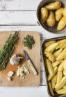 Vista superior de ingredientes para batatas de alecrim em uma tábua de corte de madeira e fatias de batata crua em um prato de assar — Fotografia de Stock