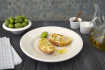 Dos rebanadas de baguette con aceite de oliva y aceitunas en plato blanco sobre superficie de madera - foto de stock