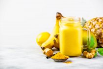 Желтый фруктовый коктейль с куркумой и ингредиентами на столе — стоковое фото