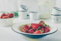 Plato con fresas frescas, tazas y leche en frasco de vidrio sobre fondo blanco - foto de stock