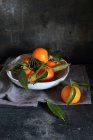 Mandarinas con hojas en tazón y sobre mesa con paño - foto de stock