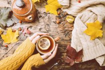 Tasse heißer Tee mit Zitrone in den Händen der Frau, die sie über dem hölzernen Herbsthintergrund hält — Stockfoto