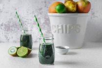 Spirulina and green fruit smoothie in glass jars, halved kiwi and limes, a big white bowl of assorted fruit on a light colored tabletop — Stock Photo