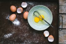 Huevos y cáscaras de huevos en bandeja para hornear, tazón con huevos líquidos y batidor - foto de stock