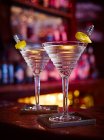 Два мартини в крученых стаканах с лимонной цедрой на металлических палочках у барной стойки — стоковое фото