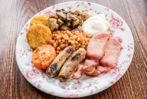 Traditionelles englisches Frühstück mit Speck, Würstchen, gegrillten Tomaten und Pilzen, gebackenen Bohnen, Kartoffelraspeln, Kuchen und Ei auf einem Holztisch — Stockfoto