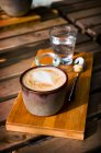 Капучіно в сільській глиняній чашці на дерев'яній дошці зі склянкою води і цукру — стокове фото