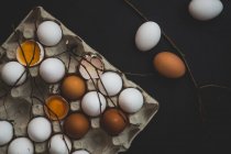 Whole and cracked open eggs in paper box and on table with tree branches — Stock Photo