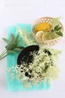 Fresh elderberry blossoms in a black bowl, with lemons in a basket on turquoise silk paper — Stock Photo