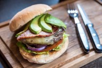 Burger mit Avocadoscheiben, Käse, Speck, Zwiebeln, Salat, Tomaten auf einem Holzbrett mit Besteck — Stockfoto