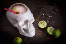 Cocktail zombie servi dans une tasse de crâne avec des pailles et des citrons verts pressés sur le fond — Photo de stock
