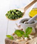 Pesto on a wooden spoon above a wooden board with basil, pine nuts and olive oil — Stock Photo
