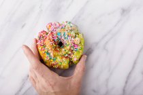 Colorful and festive unicorn donut with sprinkles on marble surface with a woman s hand holding it, unicorn food trend — Photo de stock