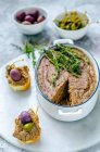 Liver pate in ceramic form with fresh thyme, and two baguette sandwiches with pate, capers and olives on a marble stand — Stock Photo