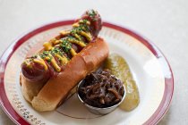 Hot dog covered in mustard and ketchup in a roll with a gherkin and onions — Stock Photo