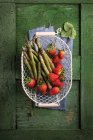 Asparagus and strawberries in a wire basket on a green wooden surface — Stock Photo