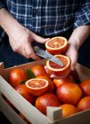 Homem cortando laranja sangue sobre caixa de madeira cheia de frutas — Fotografia de Stock