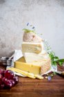 Verschiedene Käsesorten, übereinander gestapelt mit Blumen und Trauben auf hölzerner Oberfläche — Stockfoto