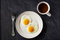 Two fried eggs with pepper in a white plate with coffee cup on black surface — Stock Photo