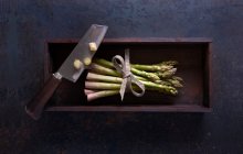 Un paquete de espárragos verdes frescos con un cuchillo en una caja de madera sobre una superficie oscura - foto de stock