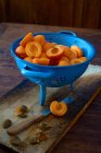 Fresh halved apricots in blue colander on wooden surface with knife — Stock Photo