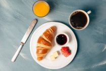 Croissant, fresh sliced strawberry, butter and jam on plate with coffee and orange juice on table — Stock Photo
