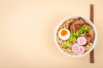 Верхній вигляд японського раму супу з локшини в білій мисці з локшиною, м'ясним бульйоном, нарізаною смаженою свининою, нарутомакі, яйце з жовткою на пастельному бежевому тлі. — стокове фото