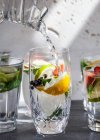 Divers verres d'eau remplis de fruits, menthe et gingembre avec de l'eau coulant du pichet dans un verre — Photo de stock