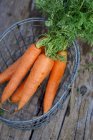 Carrots with green stems in wire basket on wooden surface — Stock Photo
