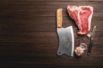 Um bife cru em T-bone com especiarias, uma faca e um cutelo numa superfície de madeira — Fotografia de Stock