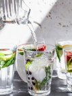 Vários copos de água cheios de frutas, hortelã e gengibre com água jorrando do jarro em um copo — Fotografia de Stock