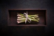 Um pacote de espargos verdes frescos em uma caixa de madeira em uma superfície escura — Fotografia de Stock