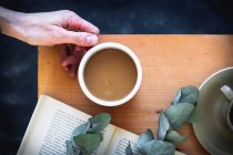 Woman's hand holding a coffee cup on a wooden table with a book — Stock Photo