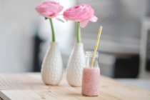 Leche de frambuesa con una paja en una botella y flores de ranúnculo rosa en dos jarrones - foto de stock