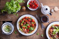 Пицца без глютена с пармской ветчиной, свежими листьями базилика и помидорами вишни подается на эмалевой тарелке, на деревянном столе — стоковое фото