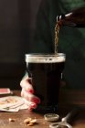 Botella de Guinness se vierte en un vaso grande que se sostiene por una mano con esmalte de uñas rojo en una mesa de madera rodeada por la tapa de la botella, abridor de botellas, nueces y cartas de juego - foto de stock