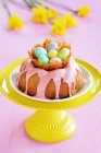 A gugelhupf with a pink sugar glaze and a caramel nest with colourful sugar eggs on a stand, with daffodils in the background — Stock Photo