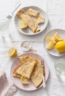Две тарелки сложенных и свернутых блинчиков с лимонными клинками на белом льняном покрытом столе и стаканах с водой — стоковое фото