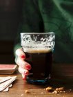 Стакан пьяного Гиннеса с женской рукой с красными ногтями и зеленым свитером, держащим стакан за деревянным столом в окружении орехов и игральных карт — стоковое фото