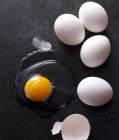 Whole eggs and raw liquid egg with broken shell and feather on black surface — Stock Photo