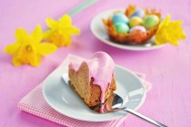 Un pedazo de gugelhupf con un glaseado de azúcar rosa, un nido de caramelo con coloridos huevos de azúcar y narcisos en el fondo - foto de stock