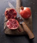 Two pomegranates, whole and halved, with a knife on an old wooden board — Stock Photo