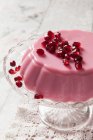 Geléia de leite rosa coberta de sementes de pomegrante em uma banca de bolo de vidro e toalha de mesa de renda vintage — Fotografia de Stock