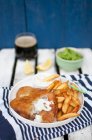 Peixe e batatas fritas, servidos com purê de ervilhas verdes, pedaços de molho de limão tártaro e cerveja escura — Fotografia de Stock