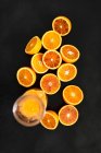 A glass jug of orange juice and halved Moro oranges against a black background — Stock Photo
